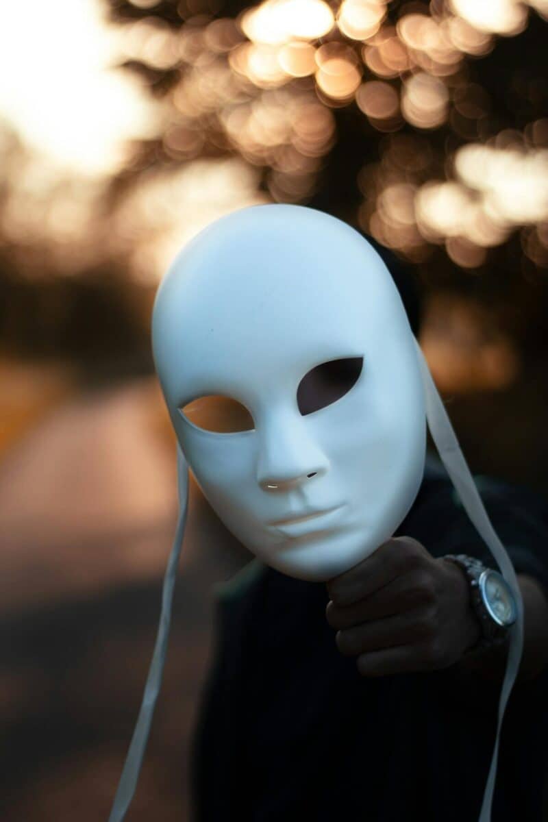 A person holds a white mask, creating a mysterious and conceptual portrait with bokeh background.
