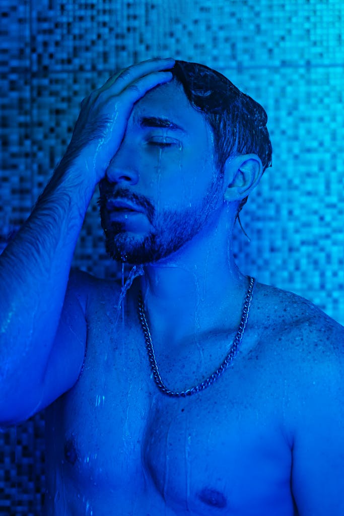 A shirtless man with eyes closed stands under blue lighting in a shower, reflecting serenity.