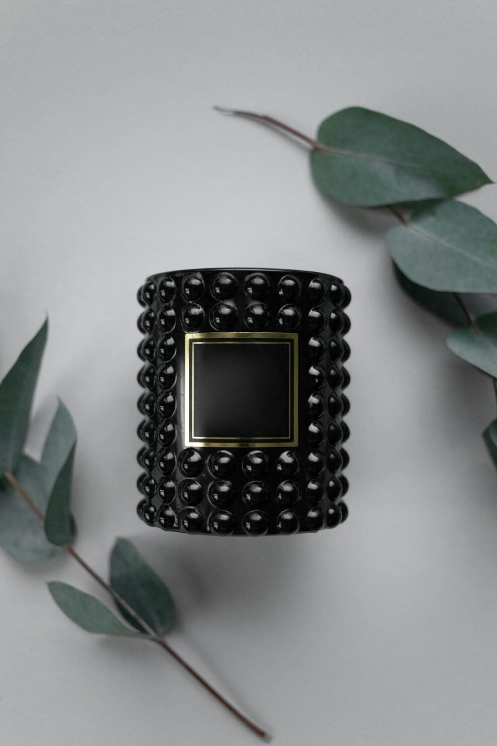 A stylish black candle with textured glass and eucalyptus leaves on a gray background.