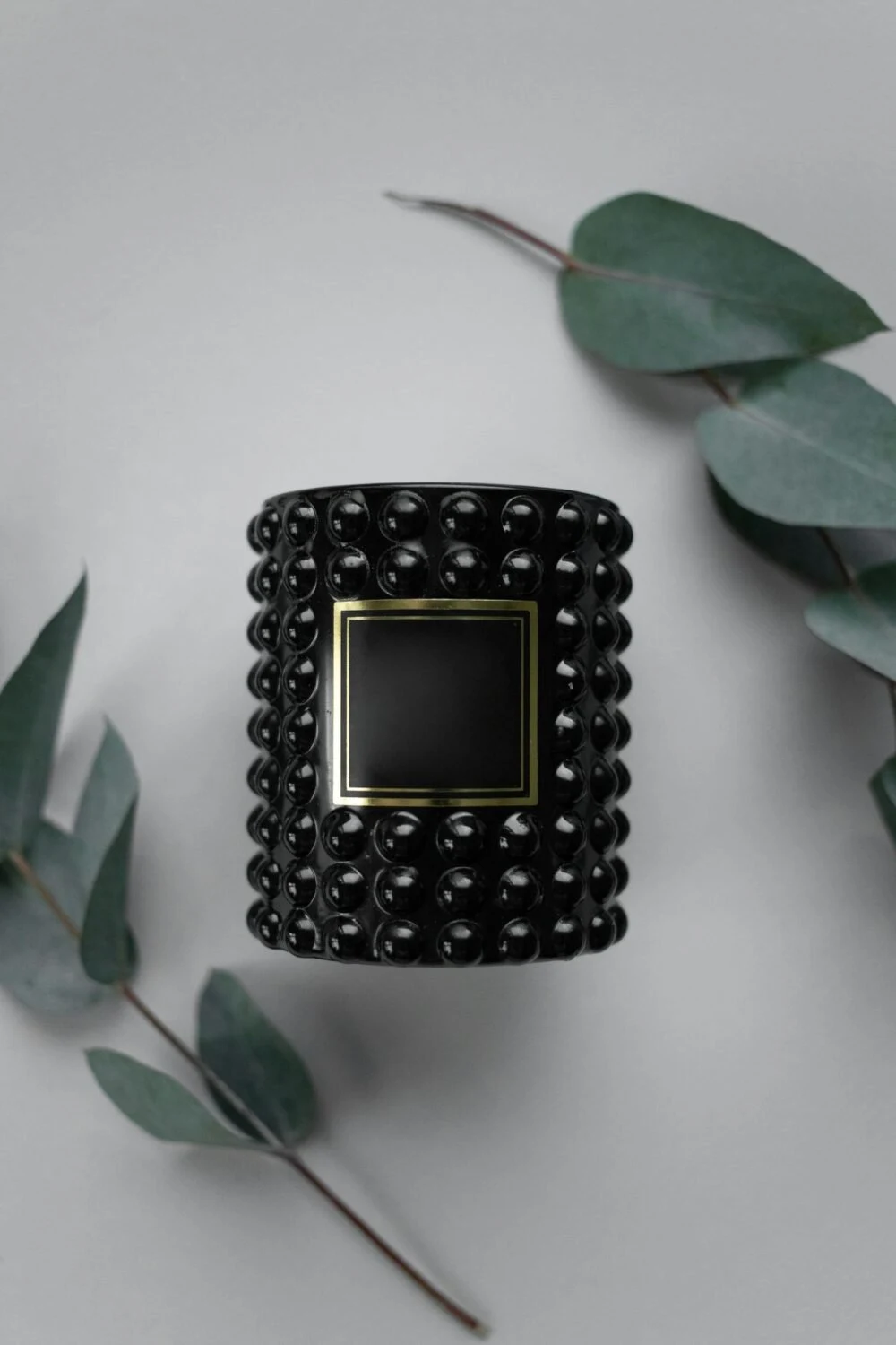 A stylish black candle with textured glass and eucalyptus leaves on a gray background.