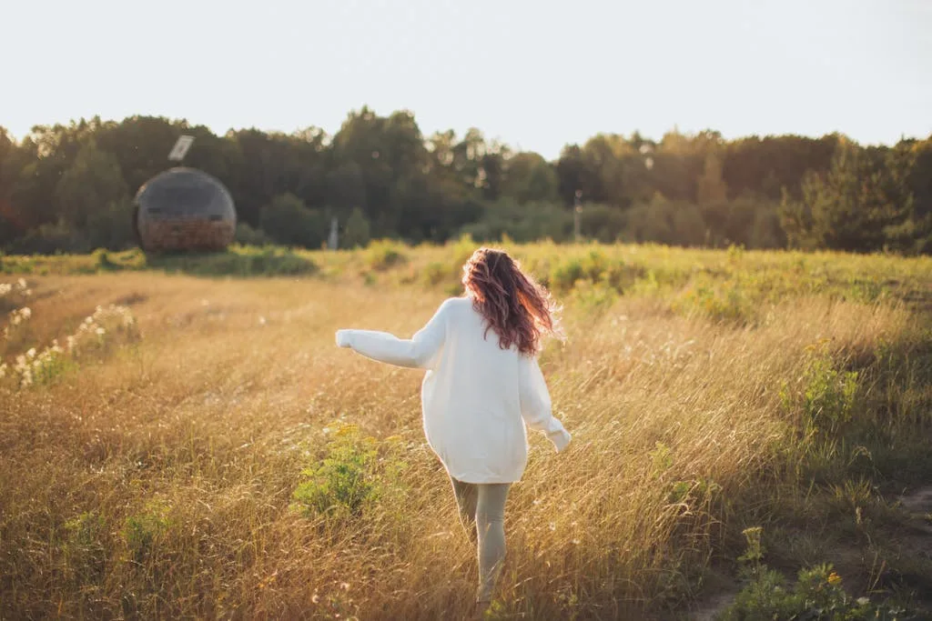 Woman in a white sweater walking through a sunlit grassy field, embracing nature and tranquility.