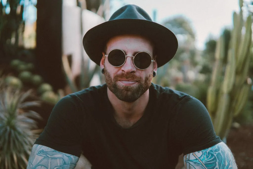 Stylish man with beard and tattoos wearing sunglasses and a hat, enjoying outdoors.