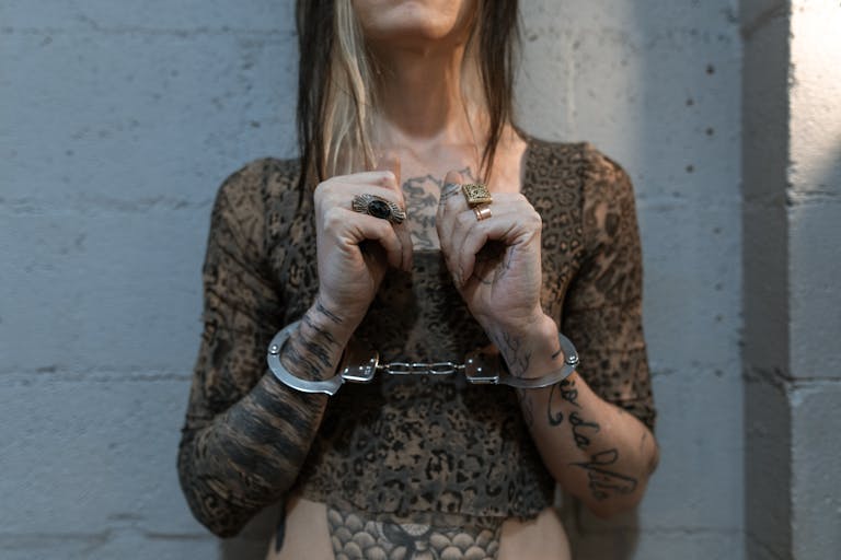 Woman with tattoos and handcuffs, standing against a brick wall, depicting crime and punishment.