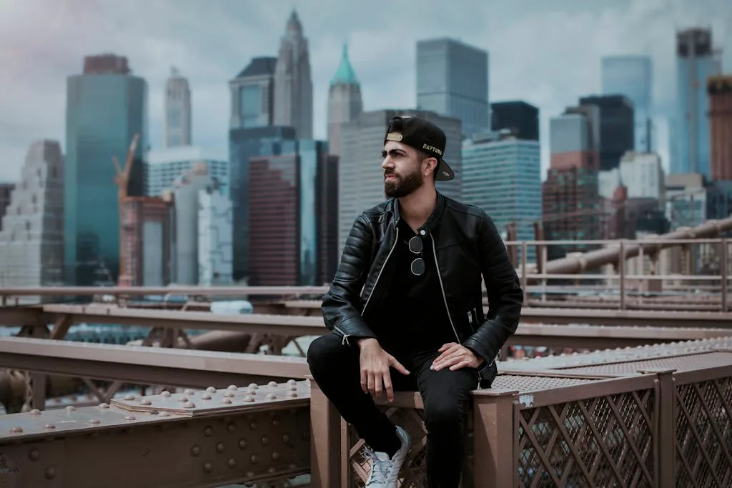 A man wearing a black leather jacket sitting on Brooklyn Bridge with New York City skyline in the background.
