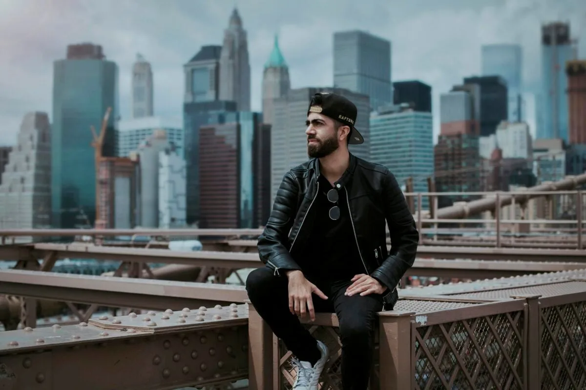 A man wearing a black leather jacket sitting on Brooklyn Bridge with New York City skyline in the background.