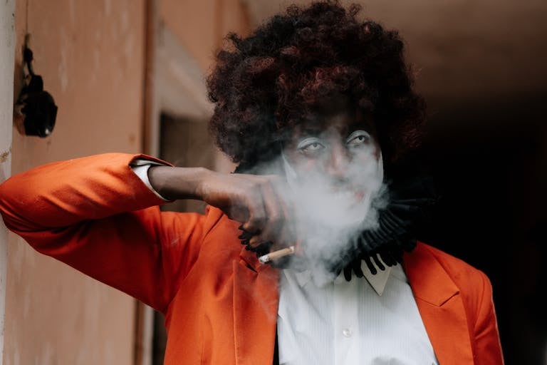 A sinister-looking clown in an orange costume smokes a cigarette indoors, exuding an eerie Halloween vibe.