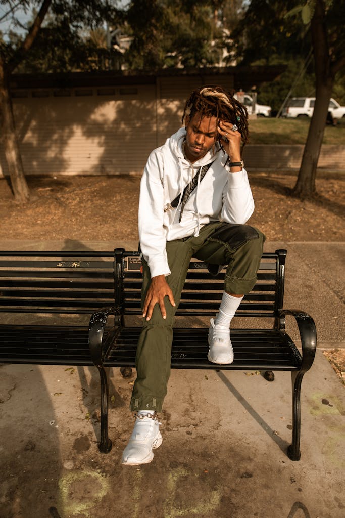 A young man with dreadlocks sits on a bench outdoors, appearing deep in thought.
