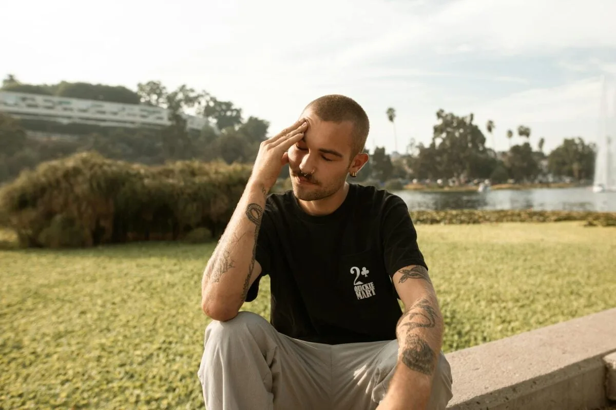 A young tattooed man sits outside, appearing thoughtful or reflective.
