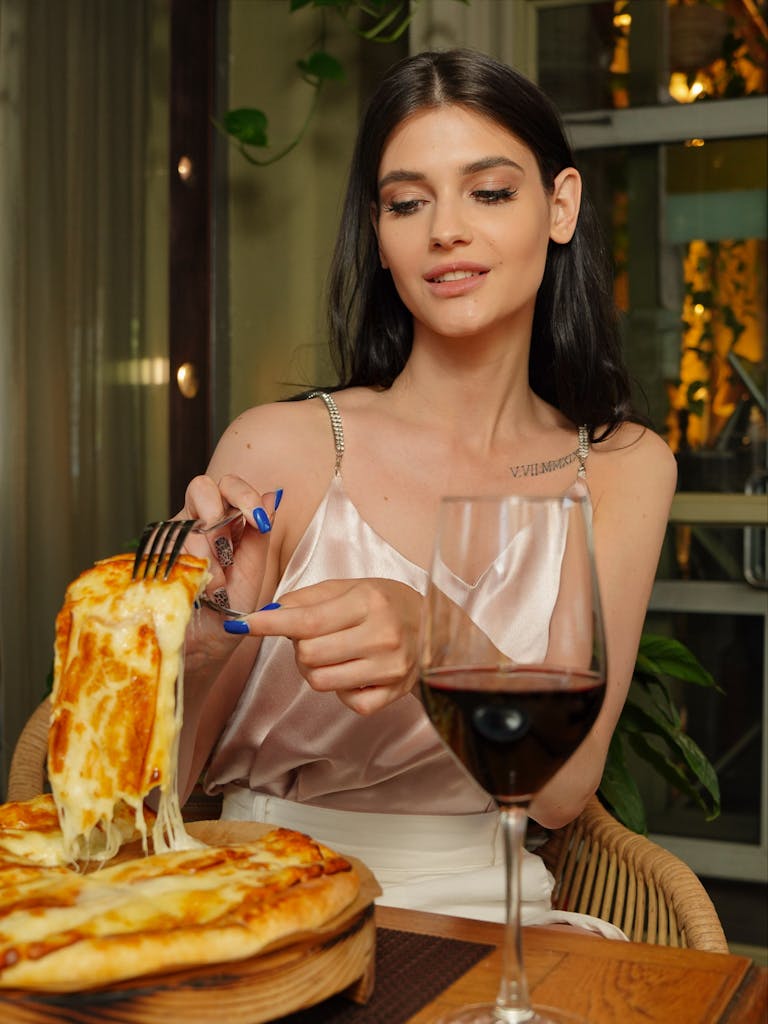 Woman with long hair indulges in cheesy pizza and red wine in an elegant setting.