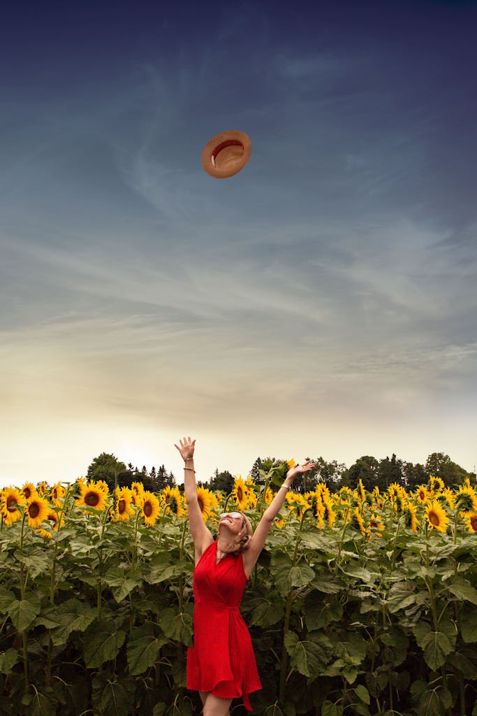 A carefree woman wearing a red dress and sunglasses joyfully throws her straw hat in a vibrant sunflower field.