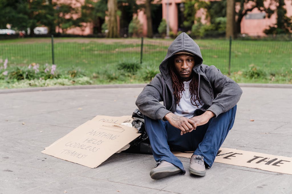 A homeless man in a city park sitting with a cardboard sign seeking help.