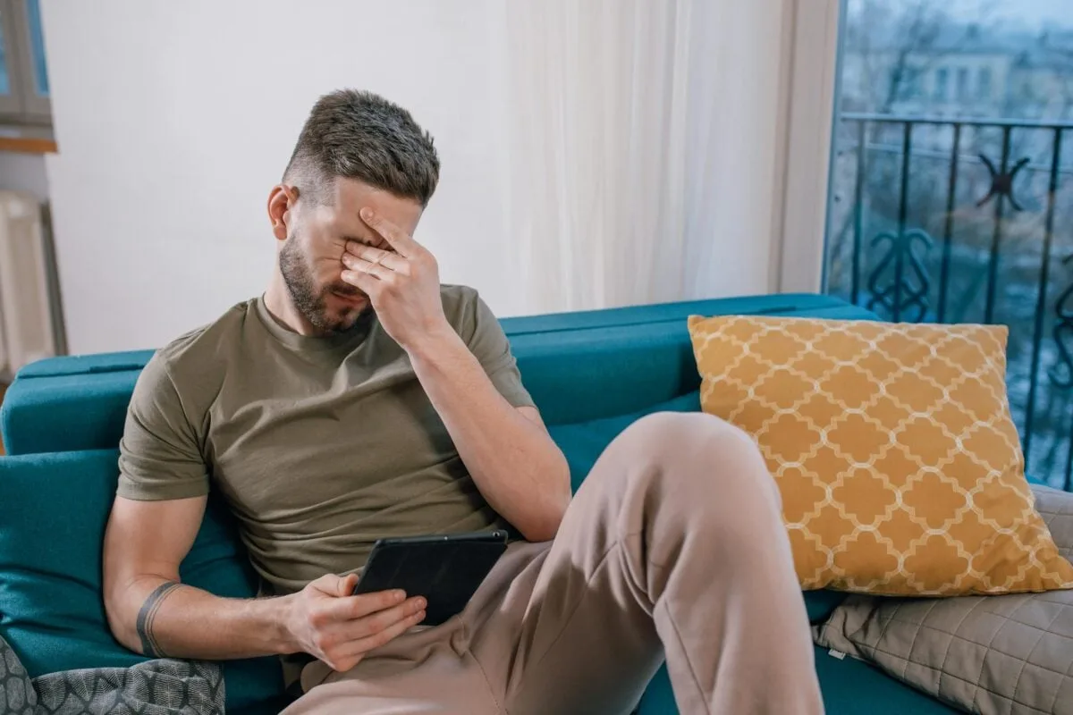 A man sits on a sofa holding a tablet, visibly stressed and covering his face with one hand.