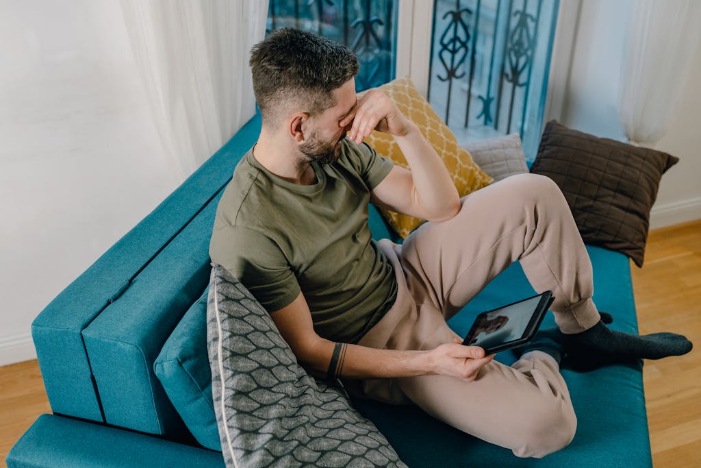 A man sitting on a sofa, looking stressed, holding a tablet inside a cozy living room.