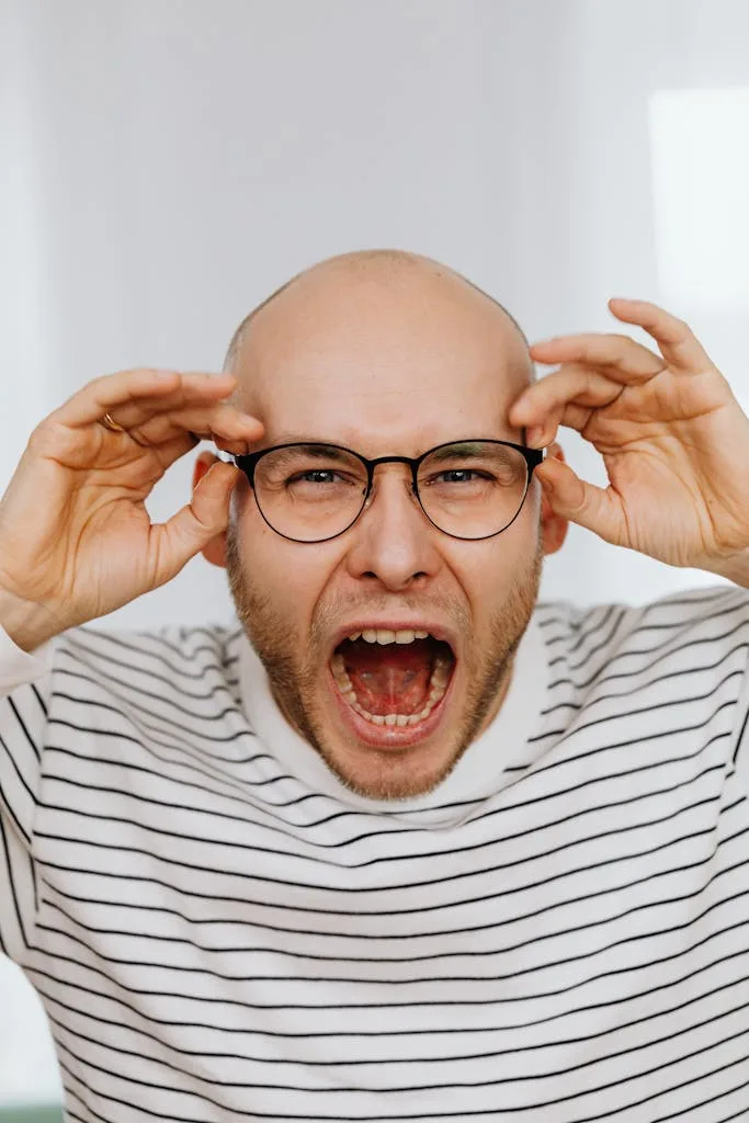 Bald man wearing striped shirt and eyeglasses, expressing anger or frustration.