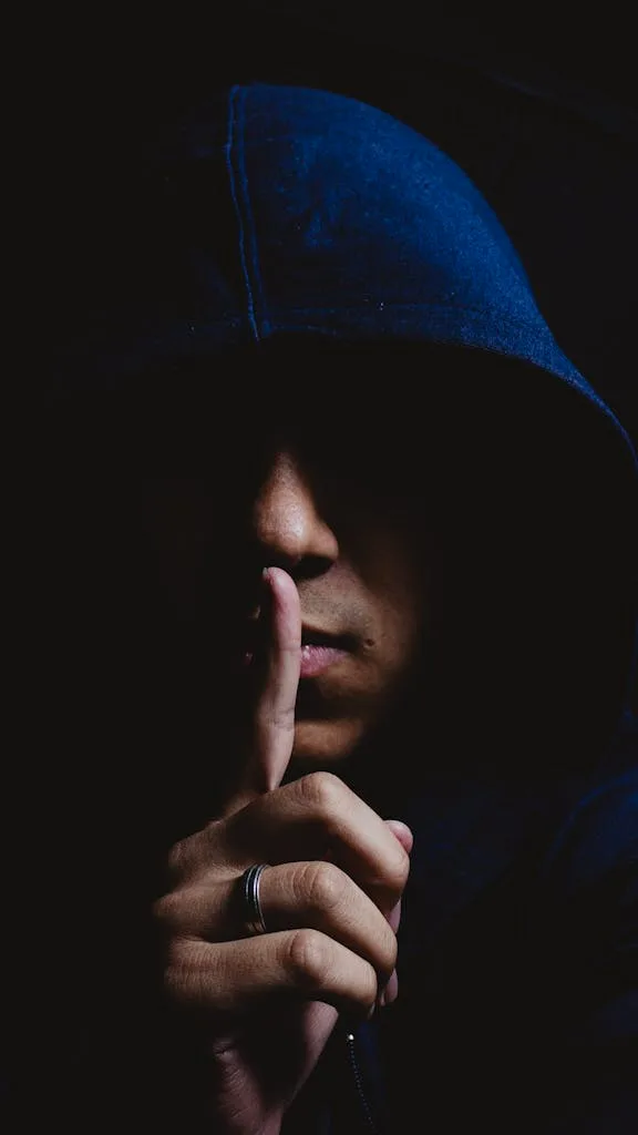 Close-up portrait of a man in a dark hoodie signaling for silence. Mysterious and moody atmosphere.
