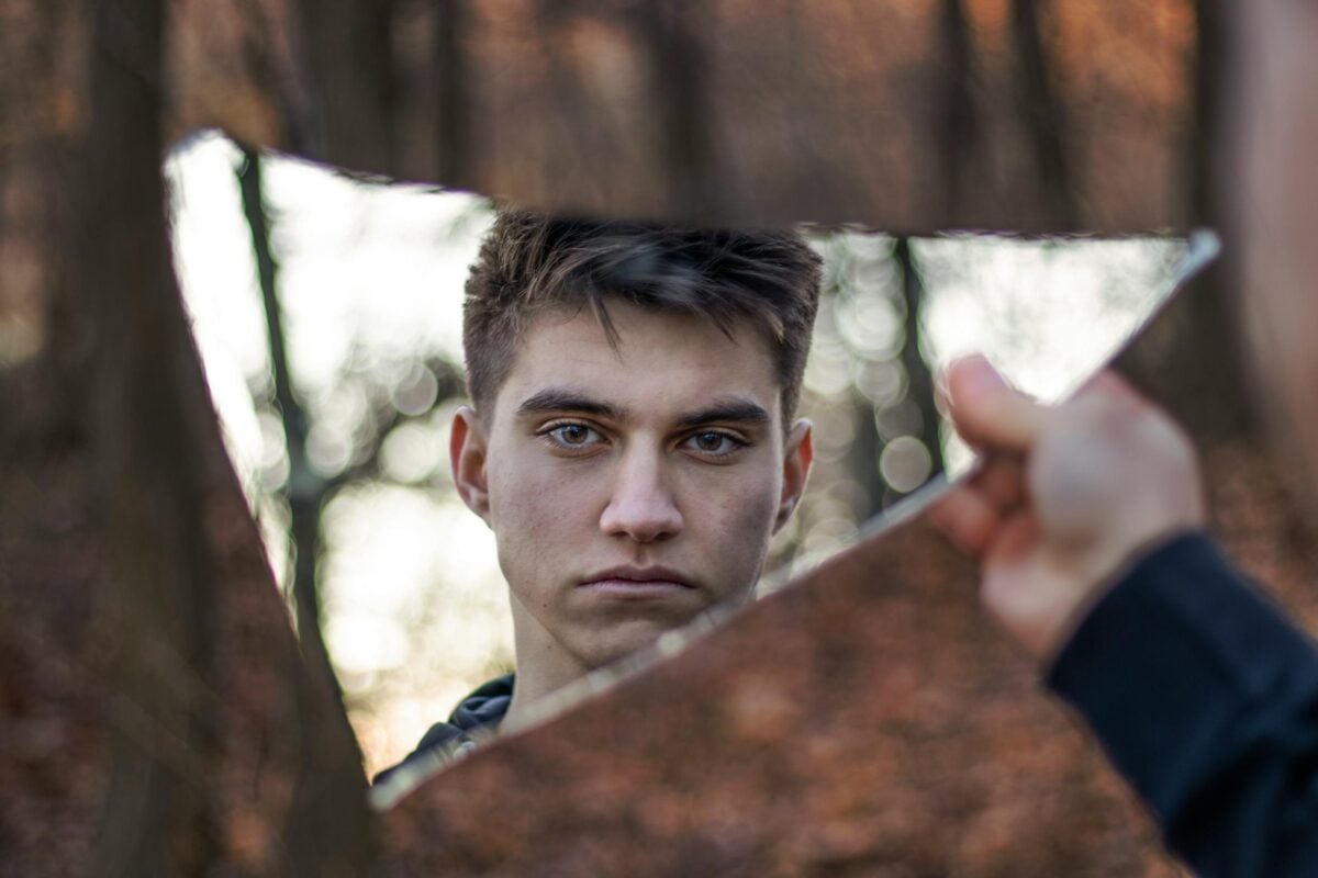A striking portrait of a man reflected in a broken mirror, set against a forest background.
