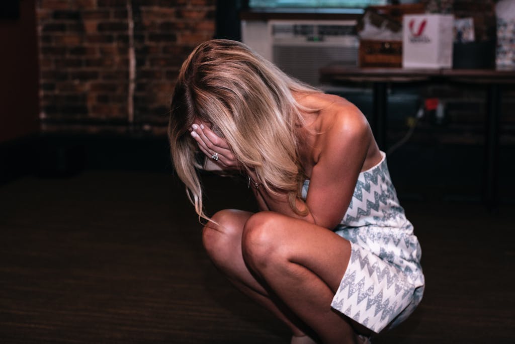 A woman in a white patterned dress sits indoors covering her face, expressing emotion.