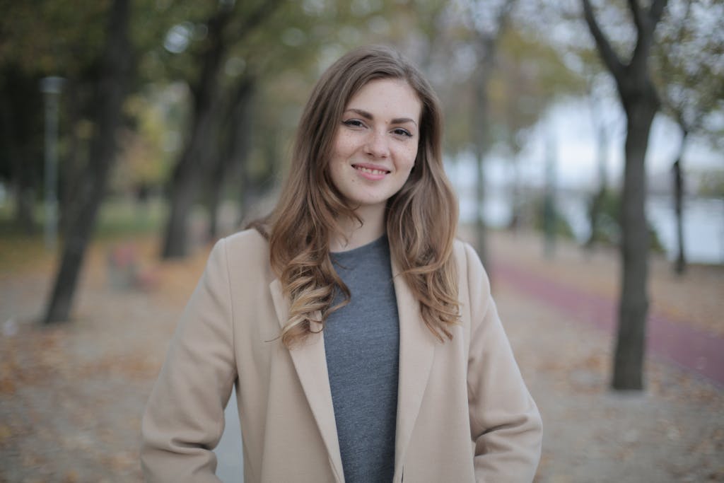 A young woman in a beige coat smiles warmly while standing outdoors during fall.