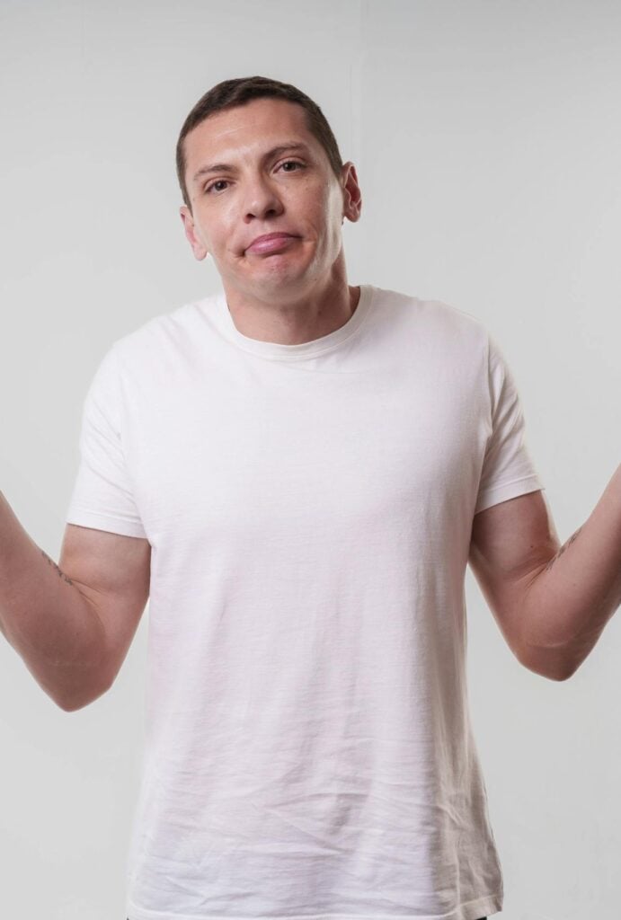 An adult man wearing a white t-shirt shrugs against a neutral background, conveying confusion or indifference.