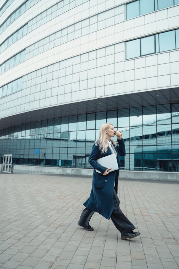 Confident businesswoman walking outside office with laptop and coffee, embracing modern lifestyle.