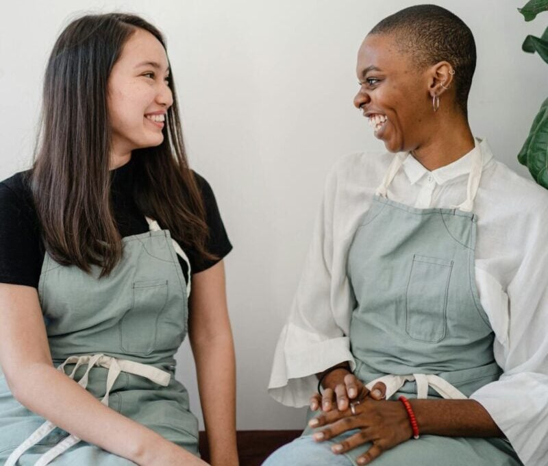 Joyful interaction between two women wearing aprons, engaging in a friendly conversation indoors.