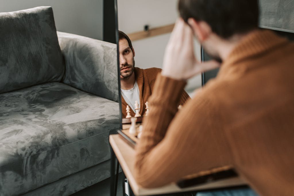 Man in brown sweater reflecting in mirror with chess pieces nearby on a calm indoor setting.