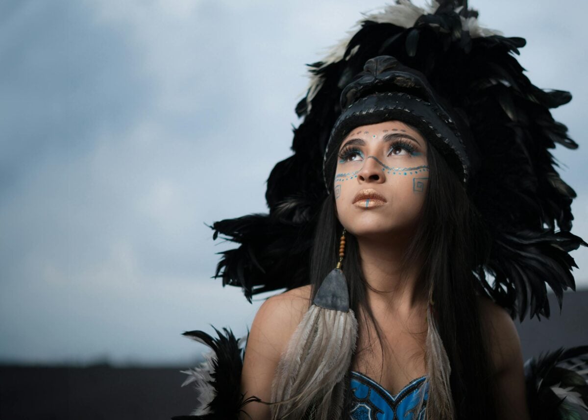 Young woman in ornate feathered headdress gazes upward with intricate face paint.