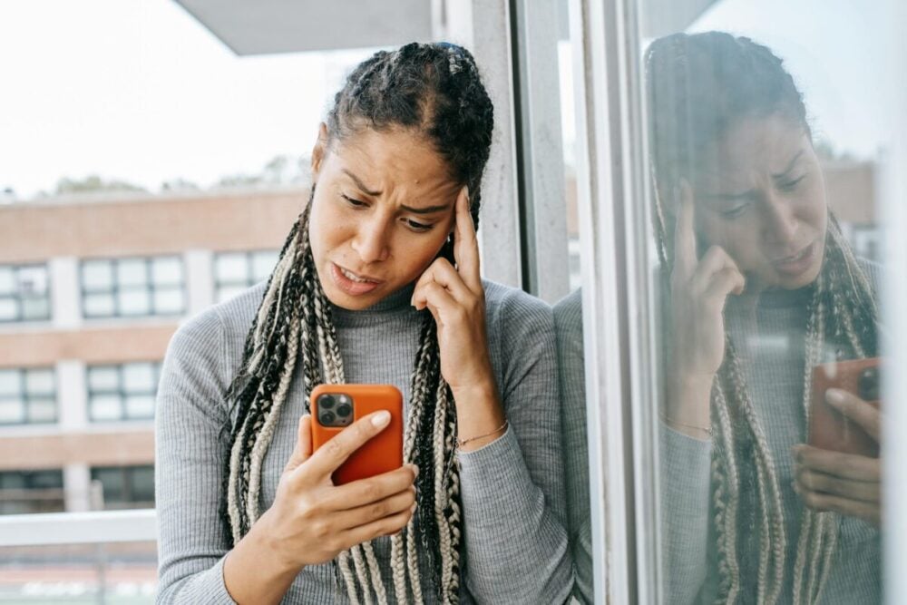 Unhappy African American female standing on balcony while holding smartphone and looking at screen