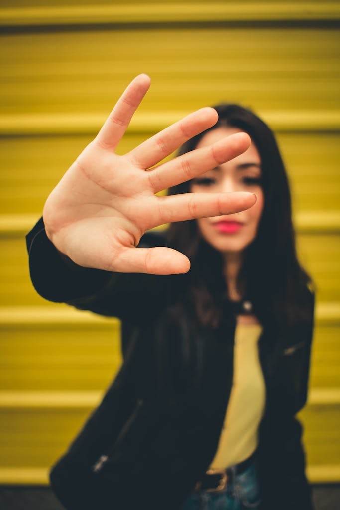 Young woman in a black jacket blocking view with hand against yellow background.