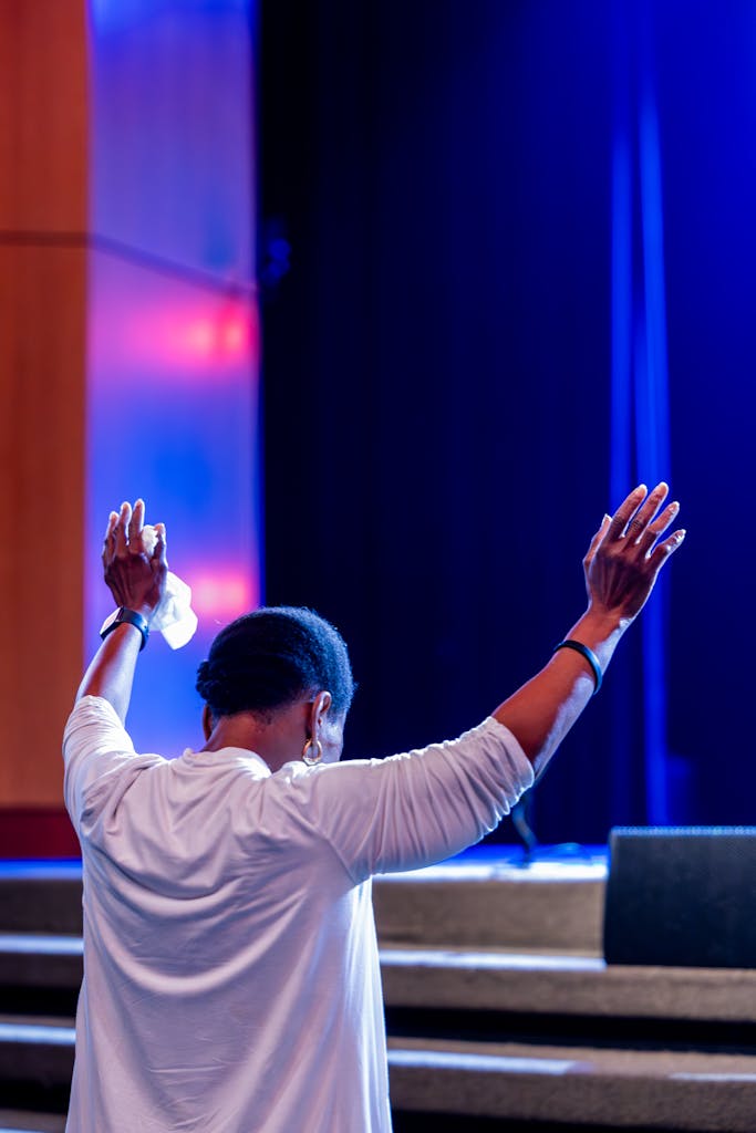 Person in prayer with raised arms on stage, captured from behind with vibrant lighting.