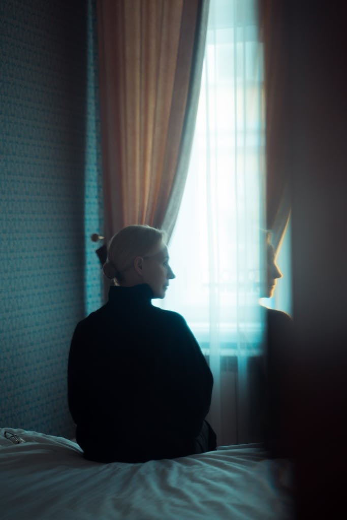 Woman sits on bed, looking out window, in dimly lit room, reflecting solitude.