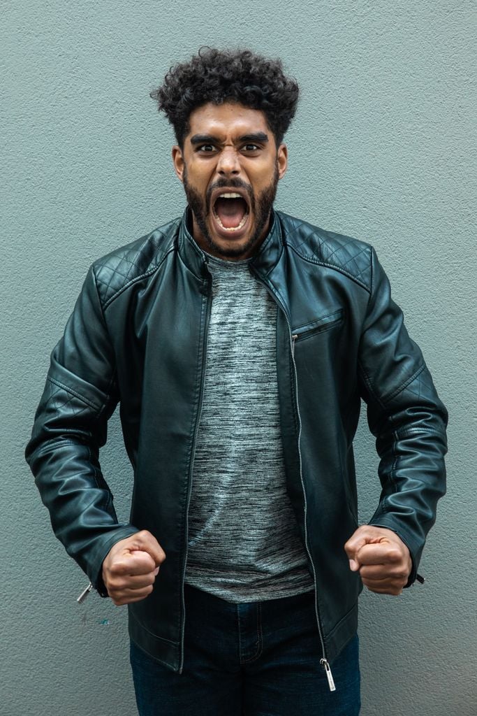 A man in a leather jacket passionately expresses anger against a plain backdrop.