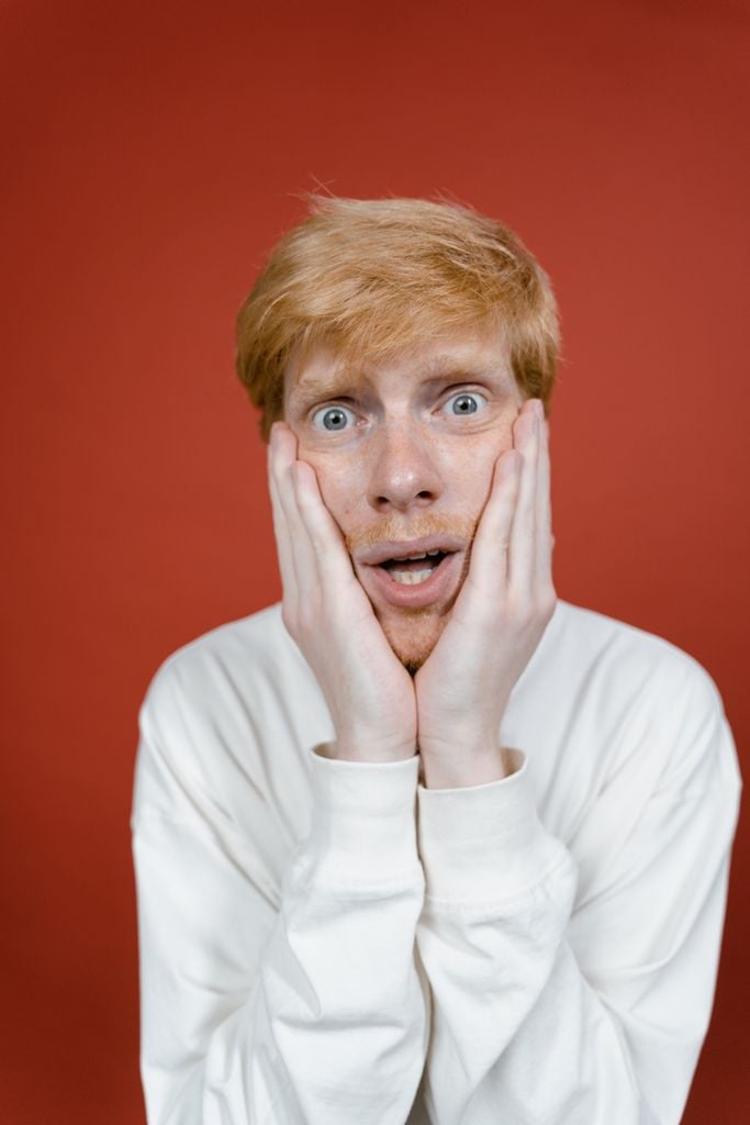 A surprised adult man with open mouth and hands on face against a red background in a studio setting.