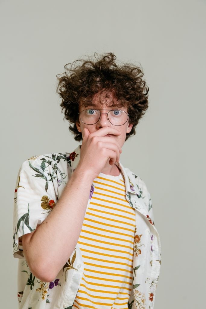 Portrait of a surprised young man wearing glasses and a floral shirt with a striped tee underneath. Neutral background.