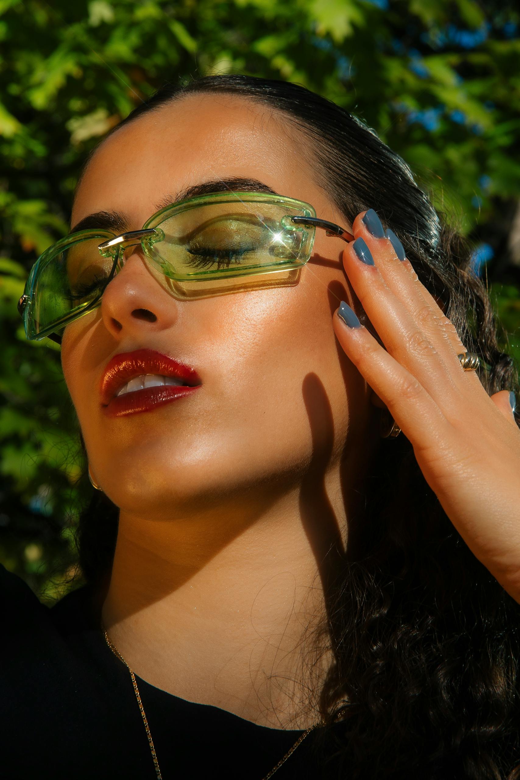 Stylish woman with green sunglasses and red lipstick posing outdoors in sunlight.
