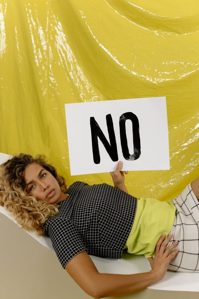 A young woman in checkered dress holds a 'No' sign, symbolizing empowerment and message advocacy in an indoor setting.