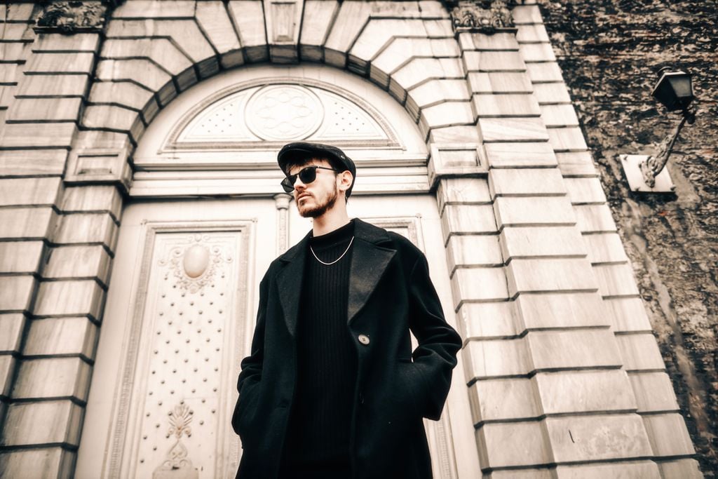A fashionable man in sunglasses and black coat standing confidently in front of a classic building.