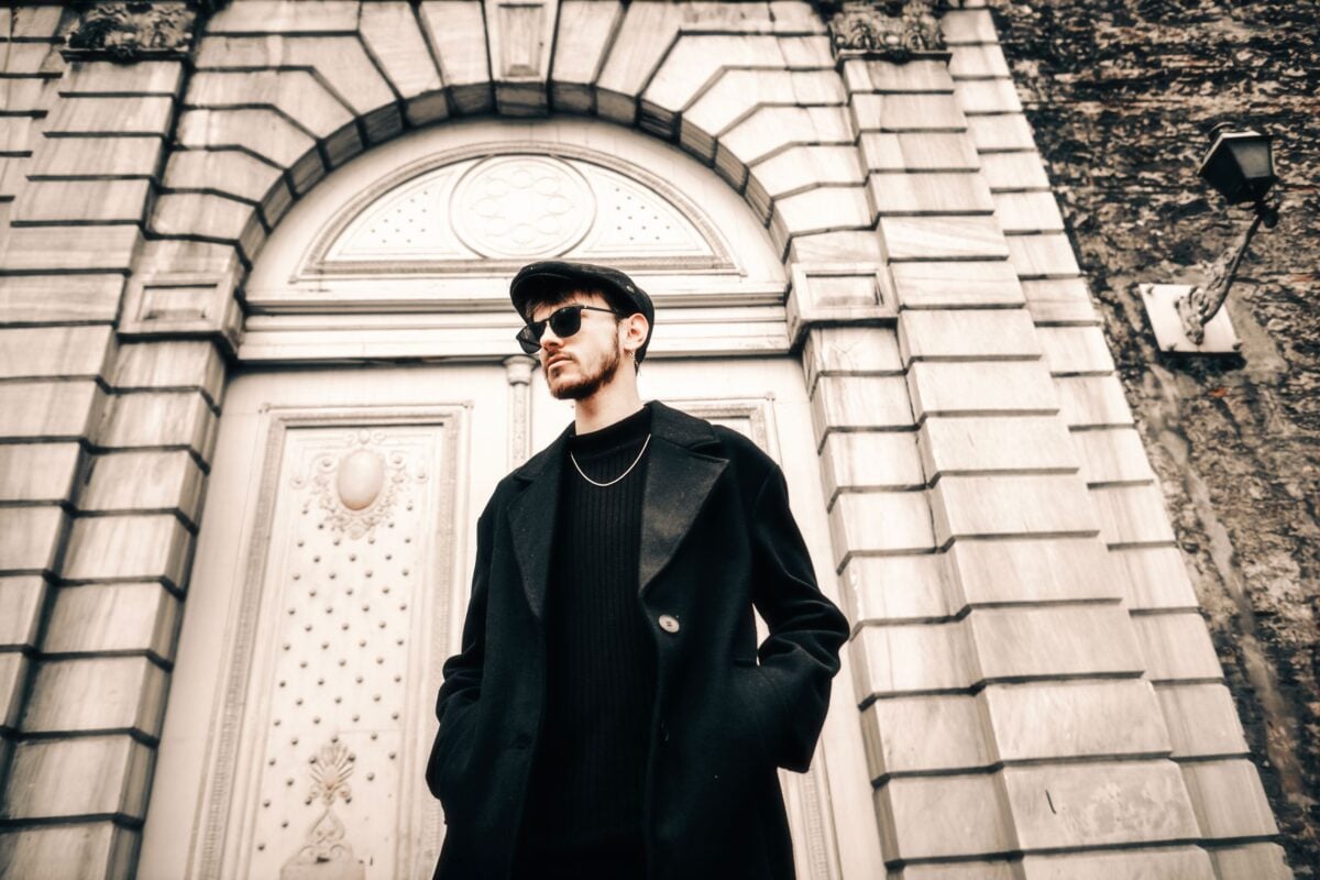 A fashionable man in sunglasses and black coat standing confidently in front of a classic building.