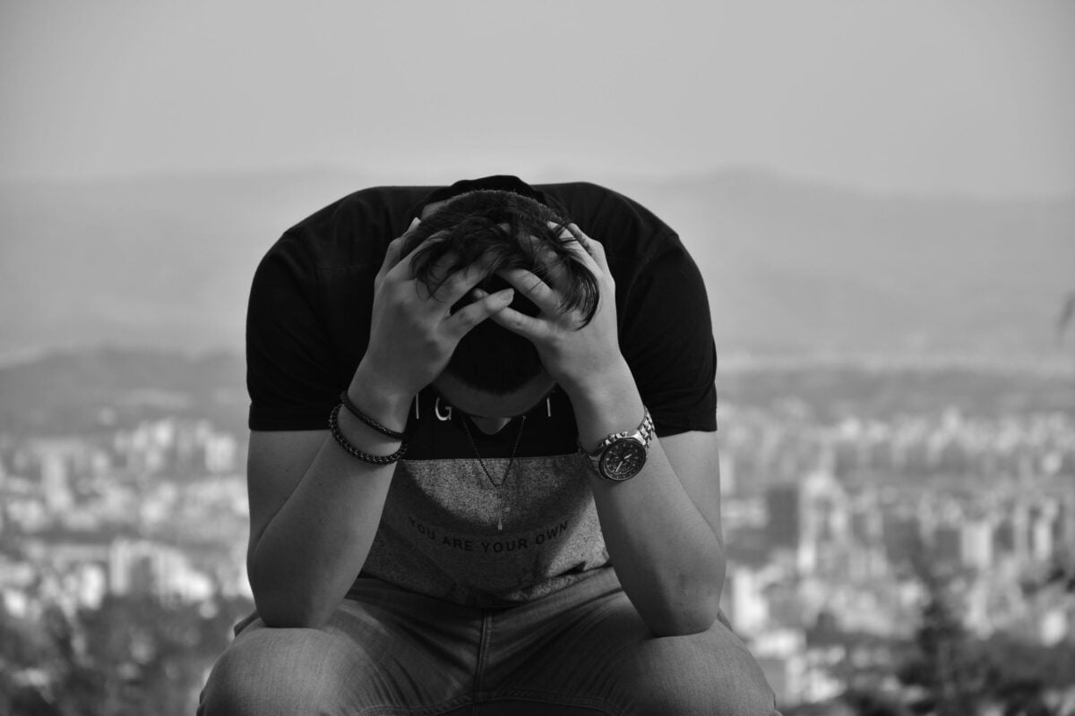 A focused young man sits with hands on head against an urban cityscape in grayscale, suggesting introspection.