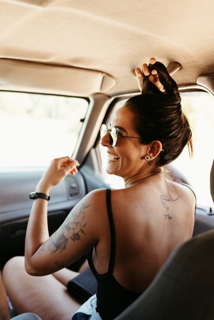 A joyful woman in a car, with sunglasses and tattoos, laughing and enjoying a summer road trip.