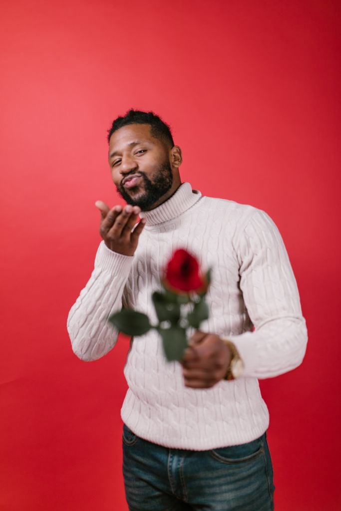 A man offers a romantic gesture with a rose and a kiss against a red background.