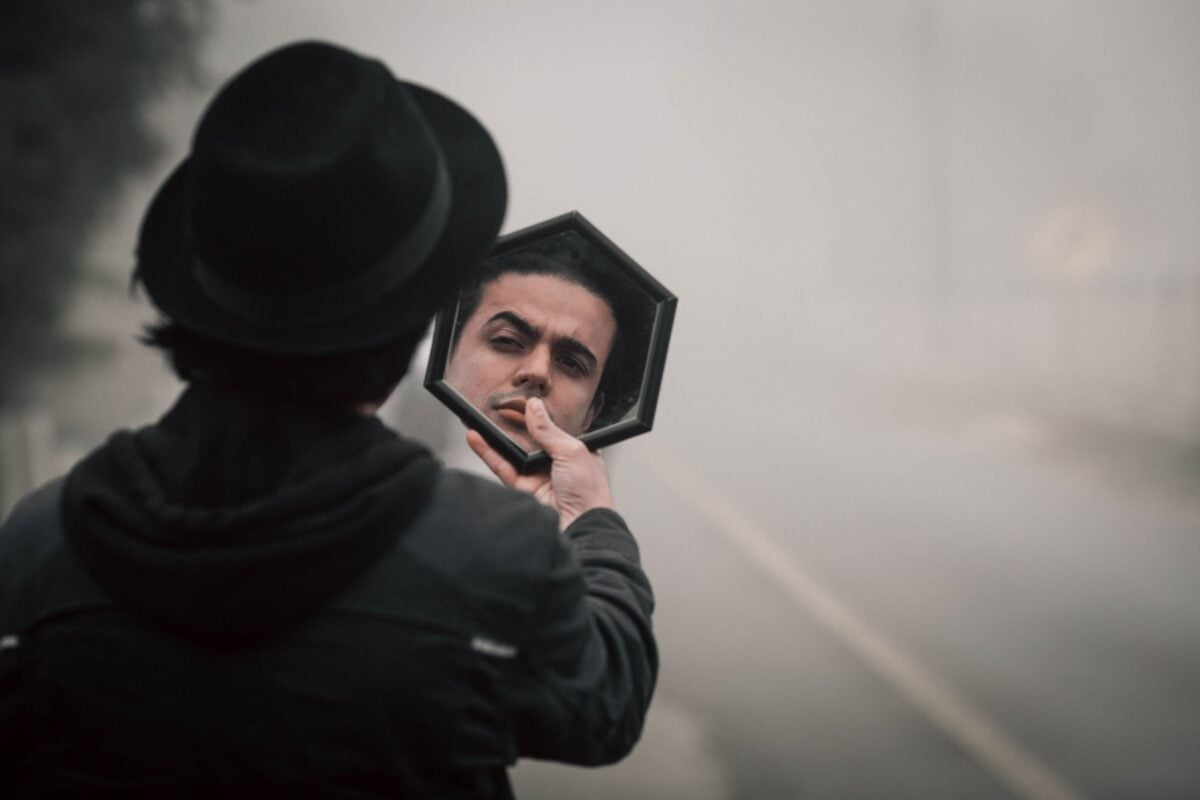 A mysterious man in a black hat reflects in a mirror on a foggy road.
