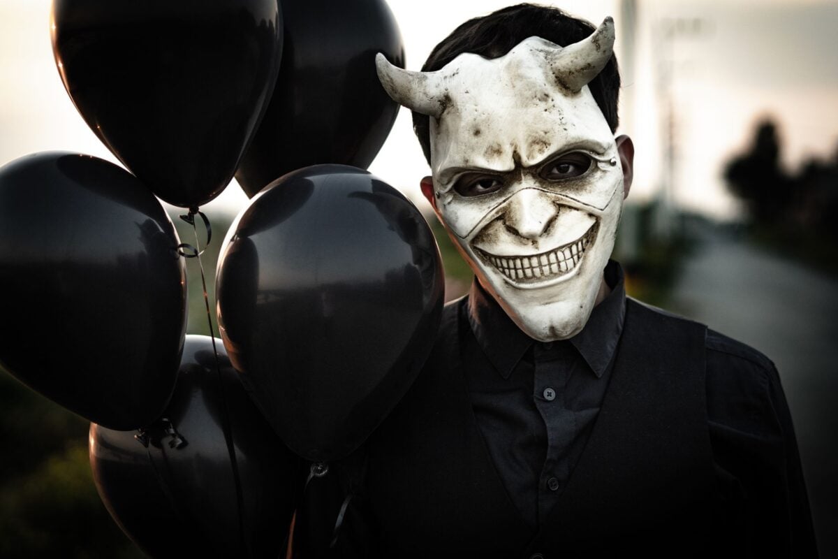 A person wearing a sinister mask holds black balloons, evoking a dramatic and eerie mood.