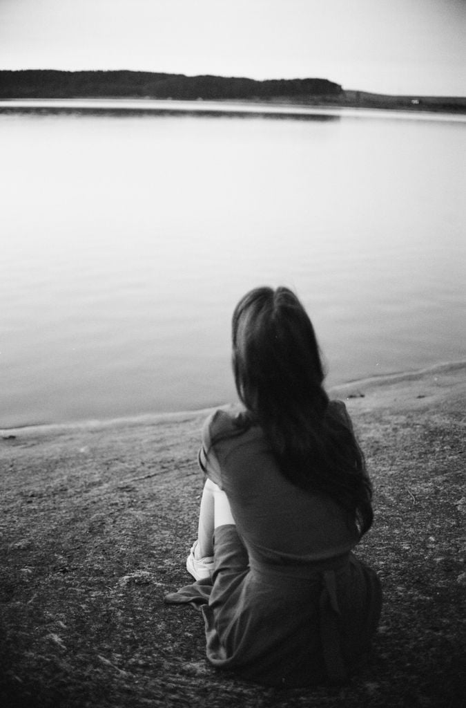 A solitary woman sits by a lakeside, reflecting in a serene black and white ambience.