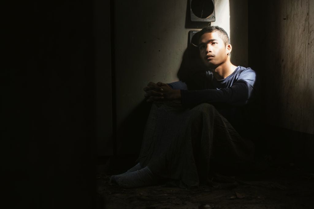 A young man sits alone in a dimly lit room, deep in thought.