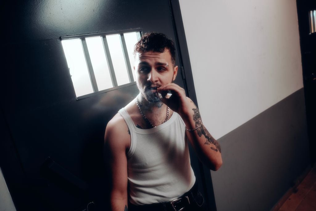 Adult man in tank top smokes cigarette near barred window, evoking prison theme.