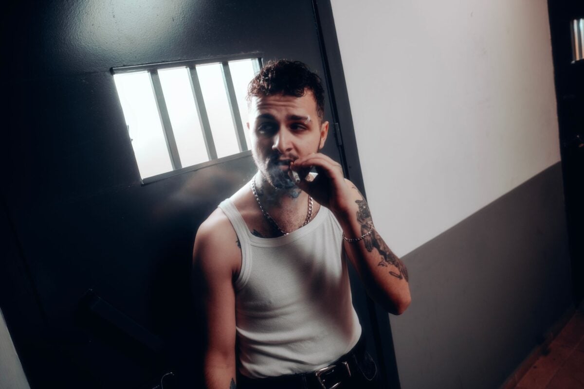 Adult man in tank top smokes cigarette near barred window, evoking prison theme.