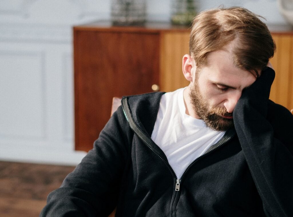 Bearded man in a black hoodie sitting in a chair indoors, appearing deep in thought.