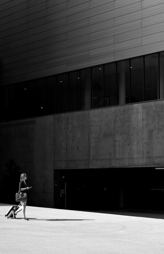 Black and white of female traveler carrying wheelie bag while walking through street to entrance of majestic gray building
