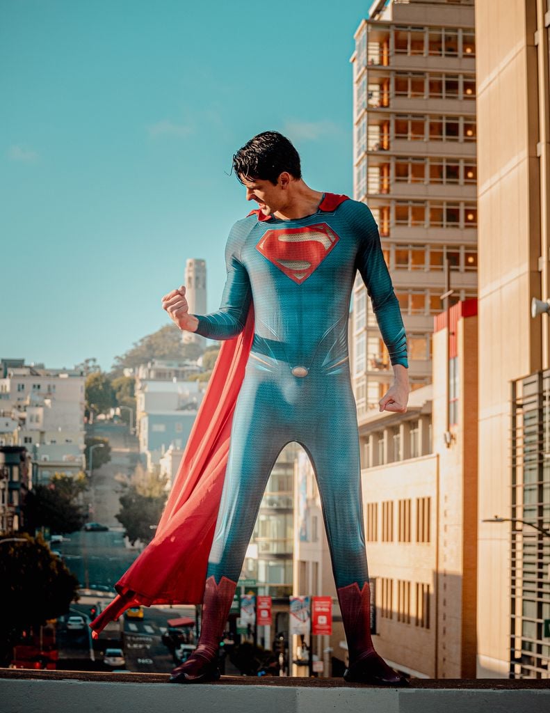 Cosplayer dressed as a superhero stands confidently in San Francisco street with cityscape backdrop.
